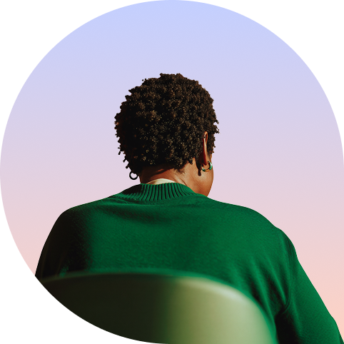 Person sedd bakifrån med naturligt lockigt hår och grön stickad tröja, fotograferad i en cirkulär ram mot en mjuk rosa-lila bakgrund.